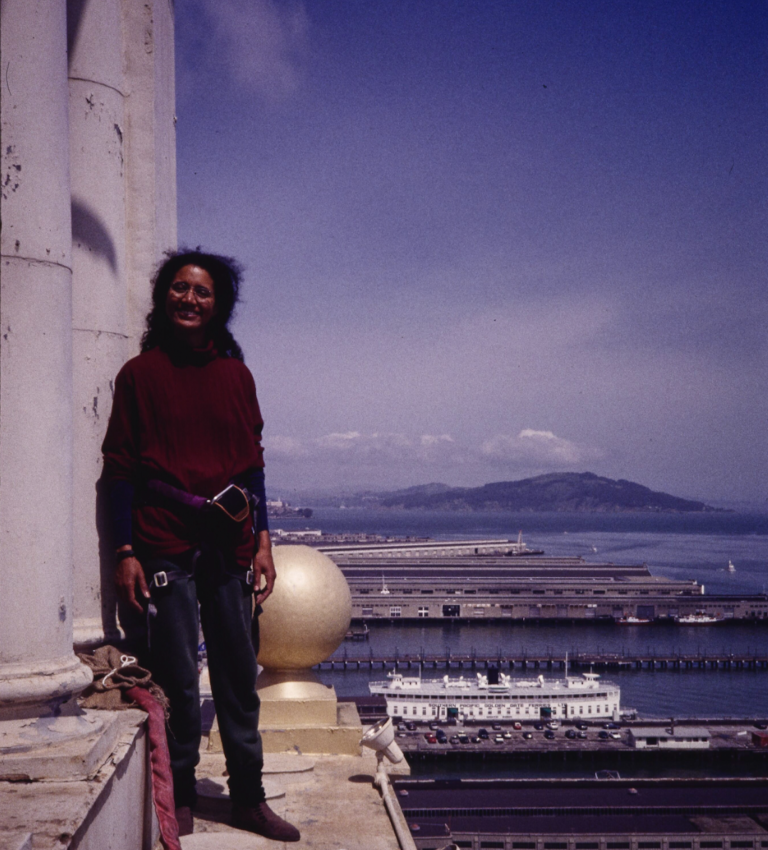 A woman in a red sweater and harness ropes stands on the edge of a giant building overlooking the San Francisco Bay.